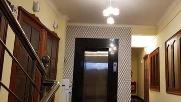 Un pasillo con un ascensor negro, una escalera con barandillas de madera y un revestimiento mural estampado.