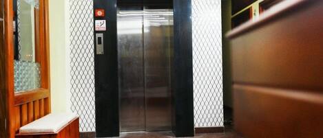 An elevator with a metallic finish, a safety sign, and a wooden bench to the left.