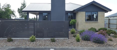 A modern two-story house with a dark exterior, a visible chimney, and a fenced front yard with gravel and small plants.