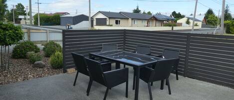 Een terras buiten met een tafel met glazen blad en zwarte stoelen, omringd door een zwart hek.