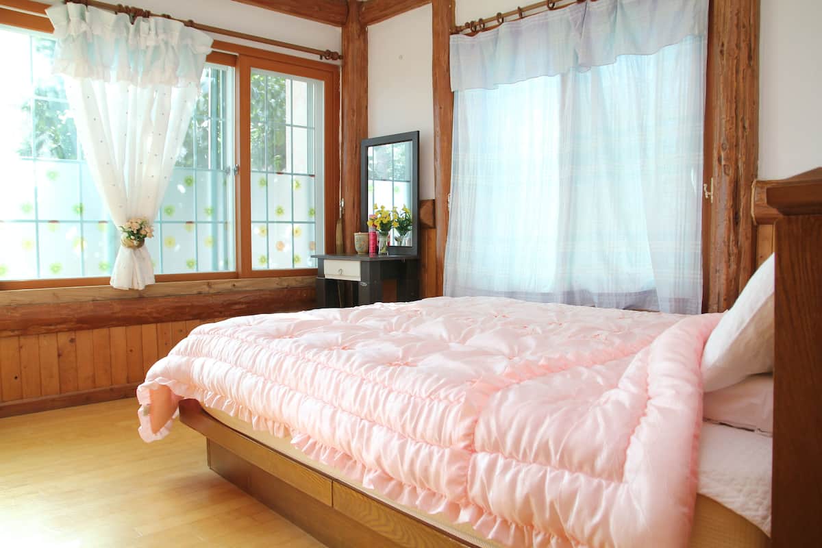 Un dormitorio con estructura de cama de madera, colcha rosa y ventana con cortina floral.
