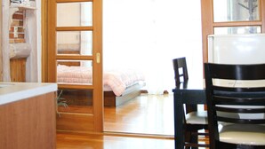 Quarto com piso de madeira, cama com cobertor rosa, porta de madeira que dá para outro cômodo e mesa com cadeiras.