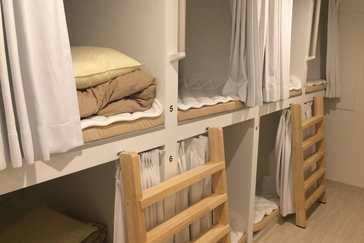 Ein Schlafraum mit Etagenbetten, weißer Bettwäsche und Vorhängen, Holzstufen und nummerierten Fächern.