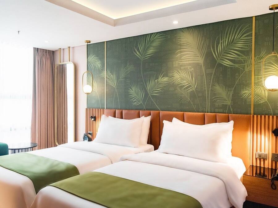 A hotel room with two beds, a green leaf-patterned headboard, a bedside table with a lamp, and a window with curtains.