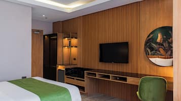 A modern hotel room with a bed, a desk with a flat-screen TV, wooden paneling, and a green chair.