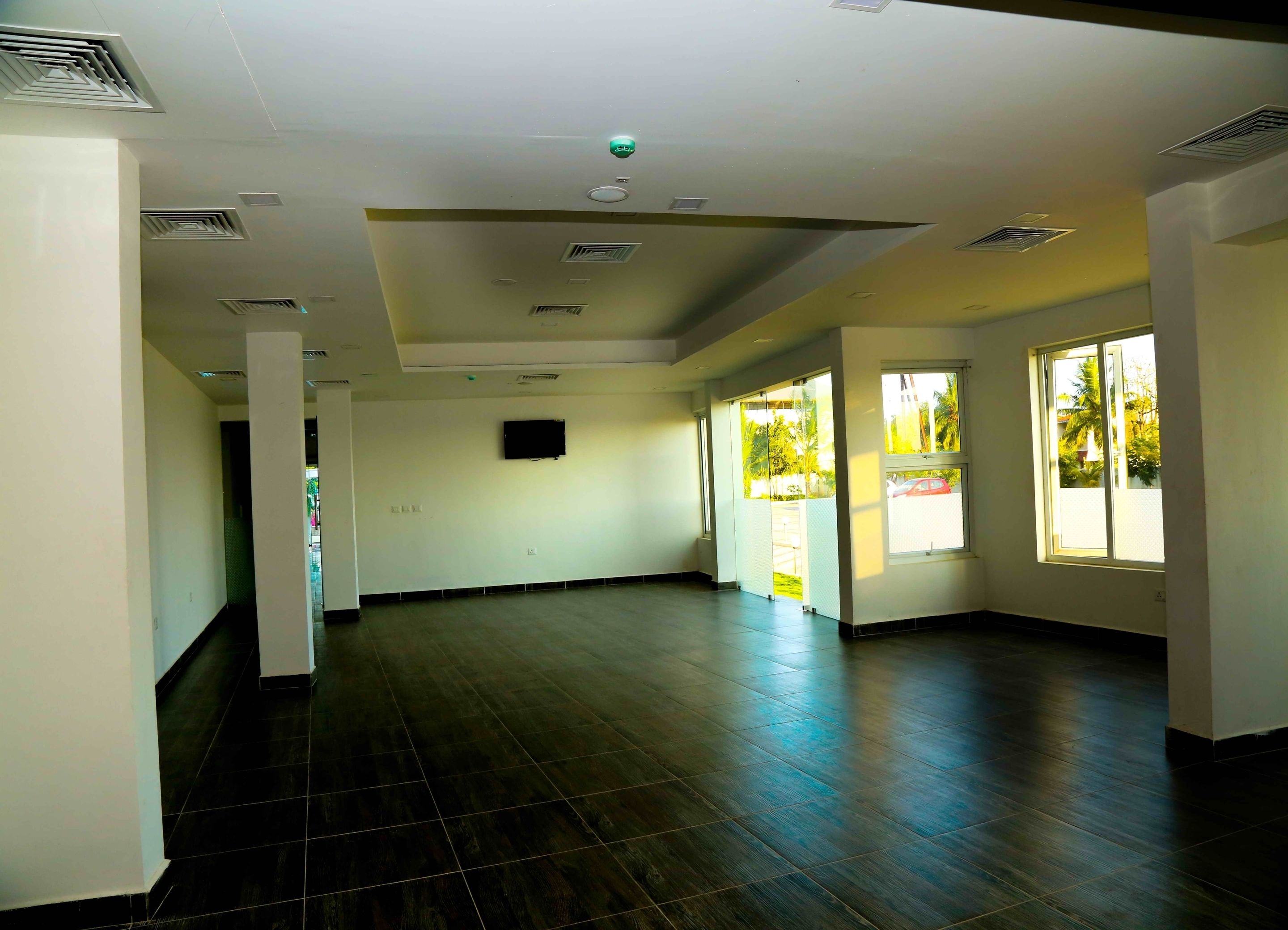 A spacious room with dark wooden flooring, white walls, and large windows allowing natural light.