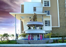 Una fuente de varios niveles con estatuas, rodeada por un borde decorado y vegetación, frente a un edificio moderno.