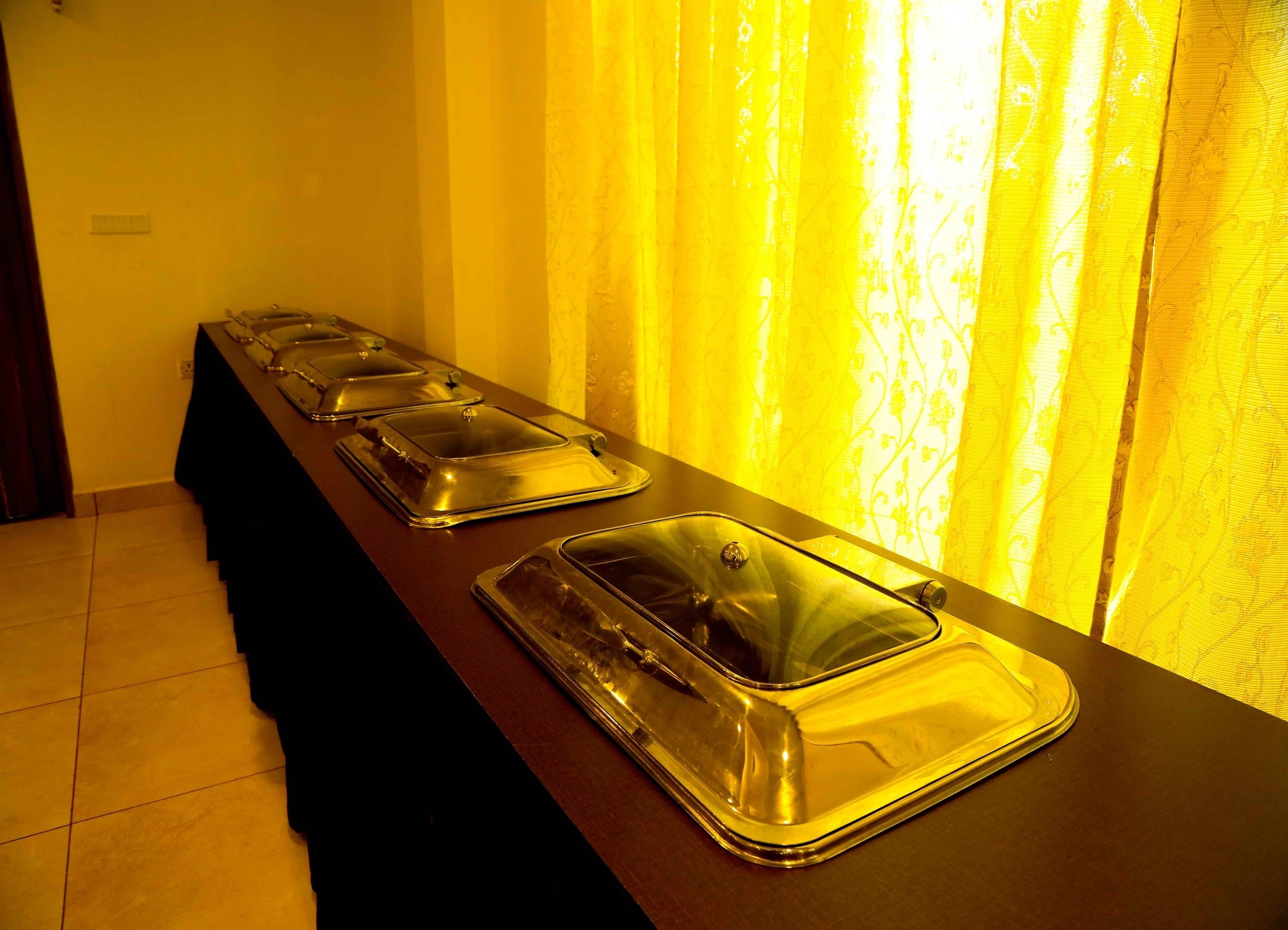 Une table de buffet avec des plats présentés sous des couvercles, un plan de travail foncé et un rideau jaune en arrière-plan.