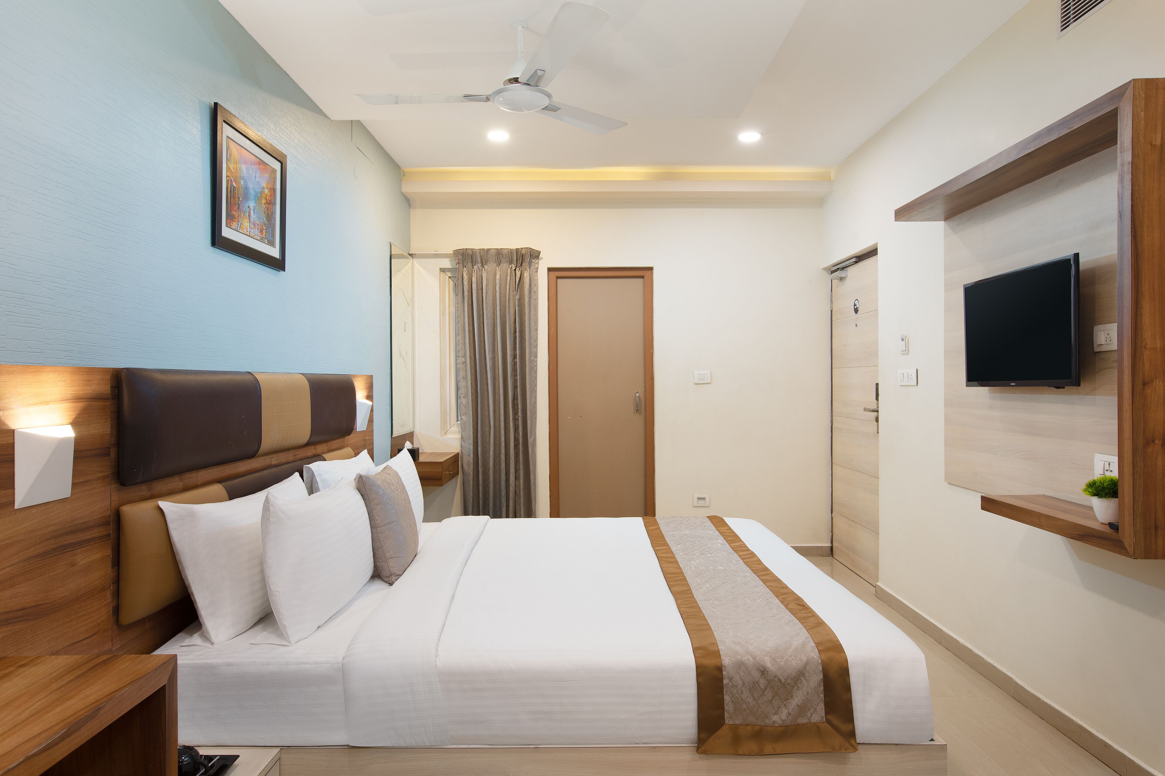A hotel room with a bed, a ceiling fan, a TV mounted on the wall, and a door leading to another room.
