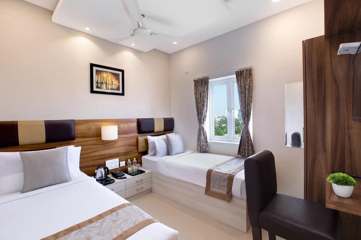 A hotel room with two beds, a desk, a chair, a window with curtains, a ceiling fan, and a wall-mounted TV.