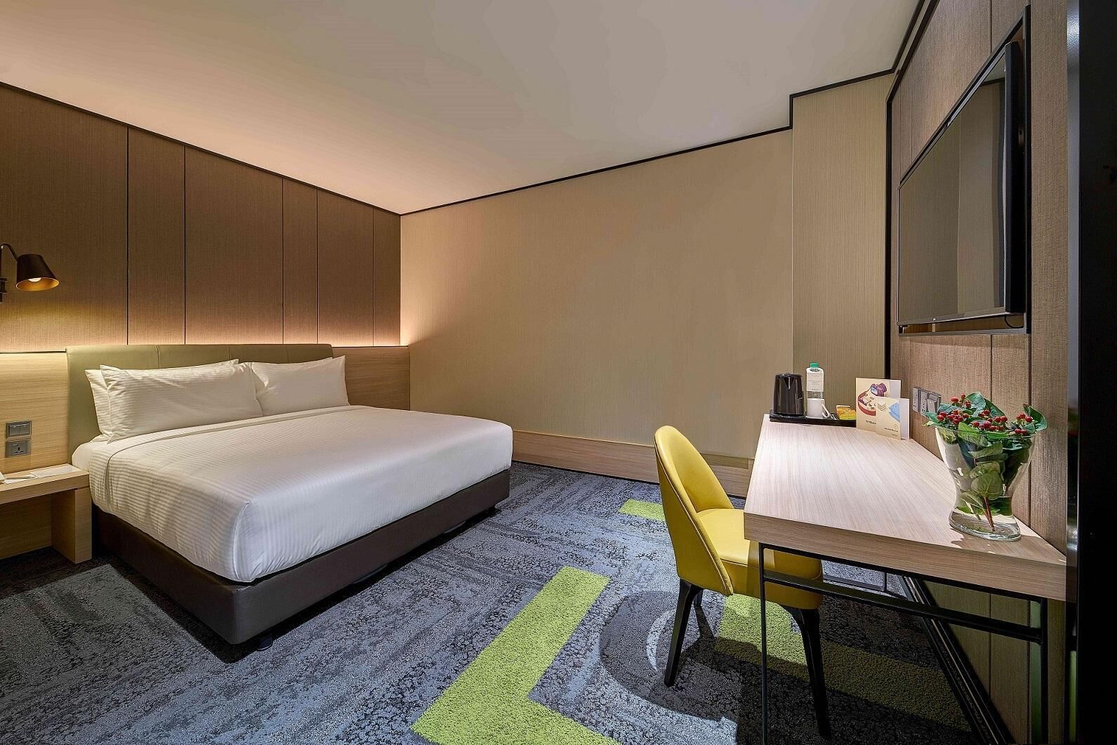 A hotel room with a bed, a desk, a chair, a TV, and a small table with a vase of flowers.