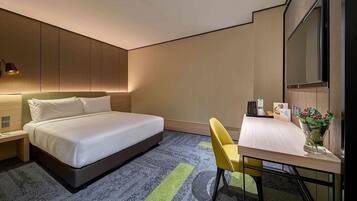 A hotel room with a bed, a desk, a chair, a TV, and a small table with a vase of flowers.
