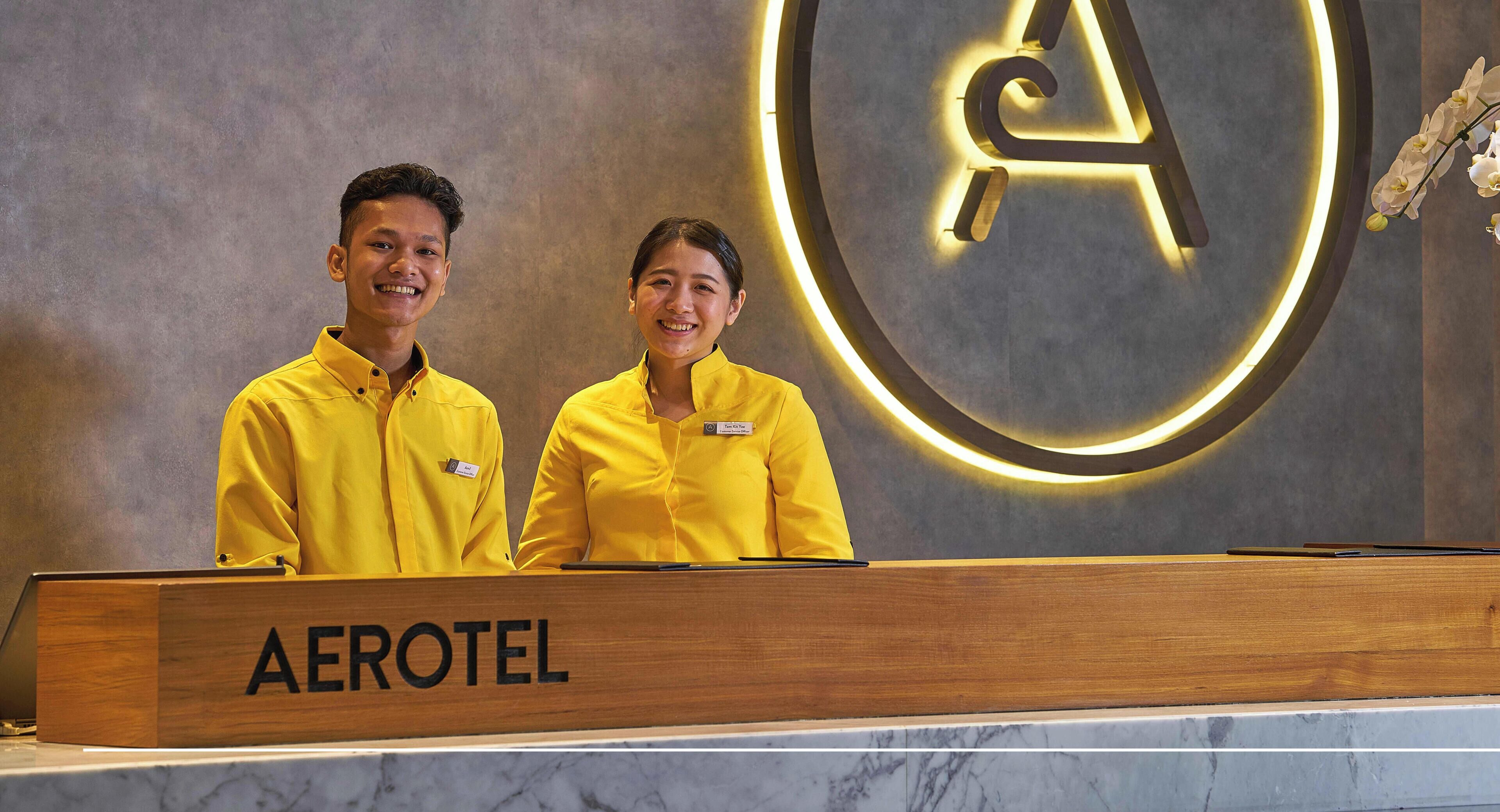 Two staff members at an Aerotel reception desk with a prominent 'AEROTEL' sign and a circular logo on the wall.