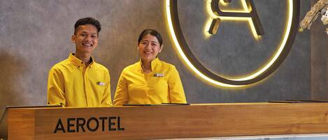 Two staff members at an Aerotel reception desk with a prominent 'AEROTEL' sign and a circular logo on the wall.