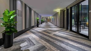 A hotel corridor with a patterned carpet, potted plants, and a reception area visible through glass doors. - Aerotel Kuala Lumpur (Sepang)
