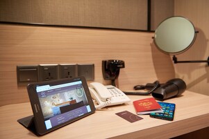 Un mostrador de hotel con una tableta que muestra un sitio web de reservas, un teléfono blanco, un teléfono negro, un espejo pequeño, un folleto rojo y un teléfono inteligente.