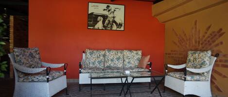 A seating area with two wicker chairs and a sofa with floral upholstery, a glass coffee table, and a red wall with a framed black and white photo.