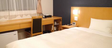 Habitación de hotel con cama, escritorio con silla, refrigerador pequeño, mesita de noche con lámpara y ventana con cortinas.