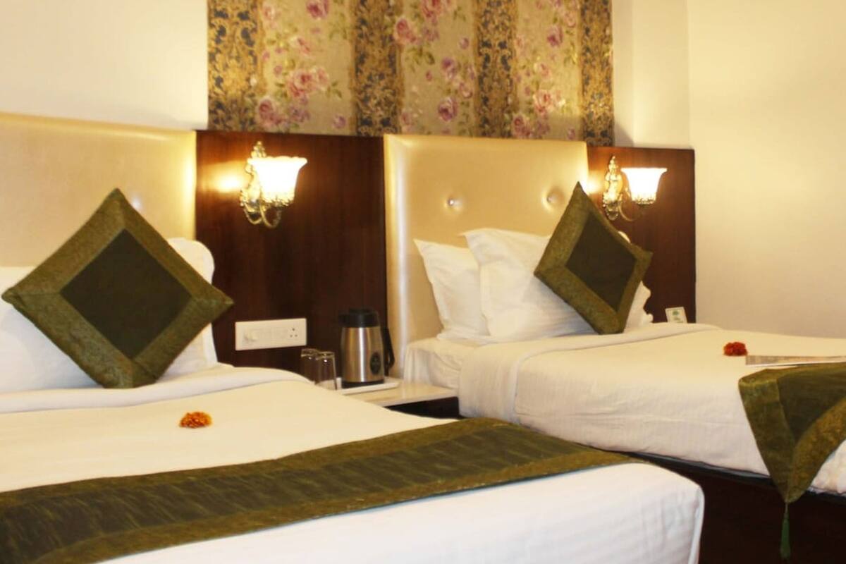 A hotel room with two beds, each with a green throw blanket, white linens, and a floral-patterned curtain.
