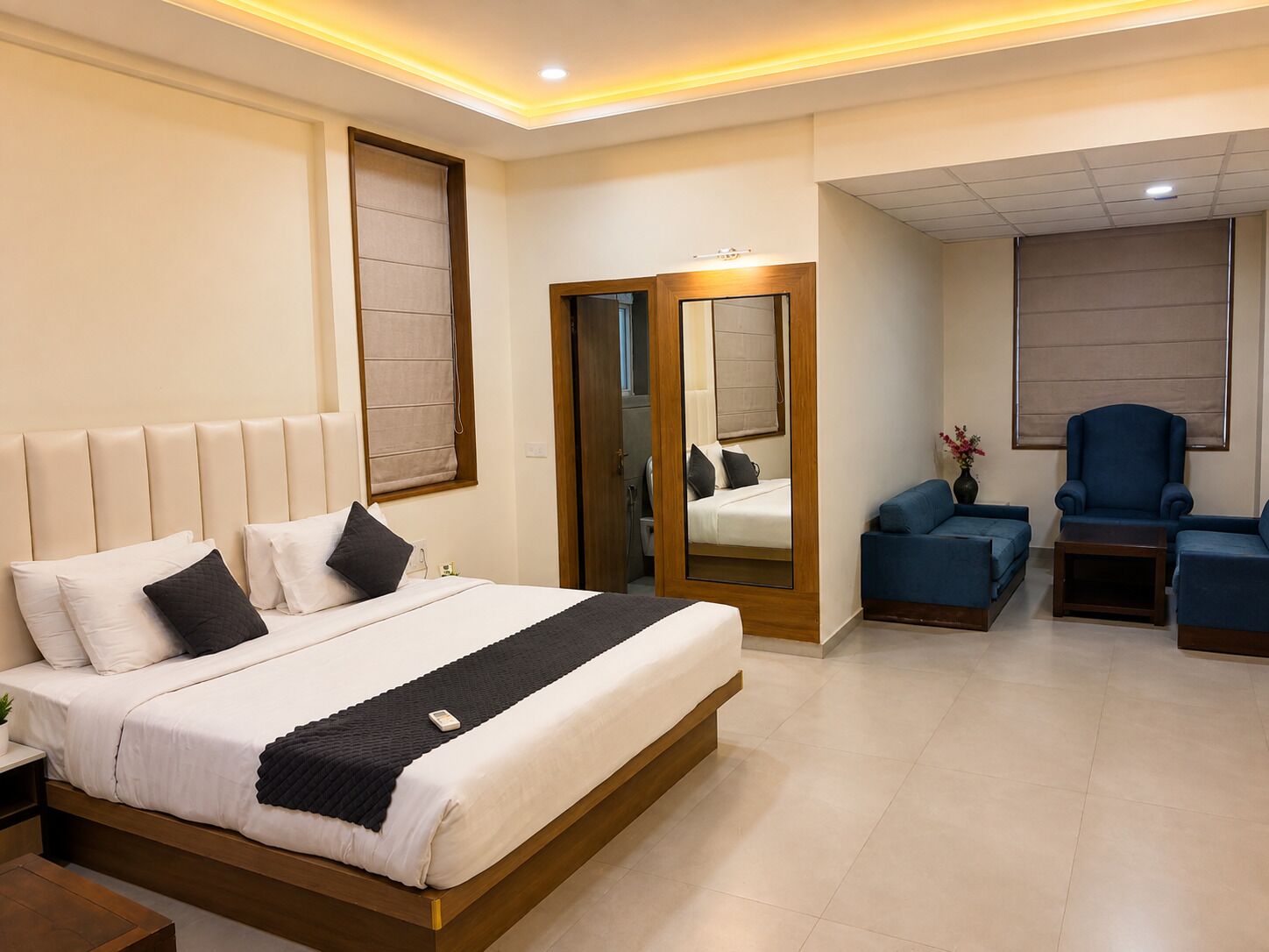 Une chambre d’hôtel moderne avec un grand lit, un coin salon doté d’un canapé bleu et de fauteuils, et un miroir qui reflète la pièce.