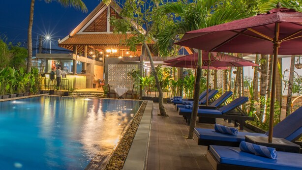 Un espace piscine agrémenté de chaises longues et de parasols, entouré de plantes tropicales et bordé d’un bâtiment dont le toit est recouvert de tuiles.