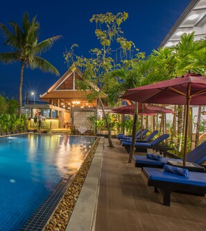 A pool area with lounge chairs and umbrellas, surrounded by tropical plants and a building with a tiled roof.