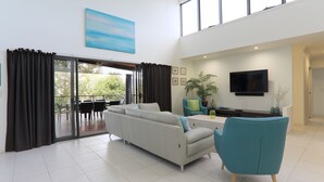 A modern living room with a sofa, a teal armchair, a flat-screen TV, and a large window with curtains. - Hilltop Views (Cannonvale)