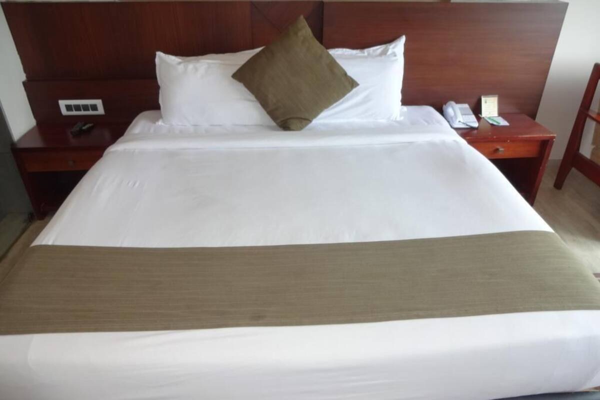 Cama bem arrumada com lençóis brancos e manta marrom, cabeceira de madeira, mesa de cabeceira com telefone e luminária, e mesa lateral com controle remoto.