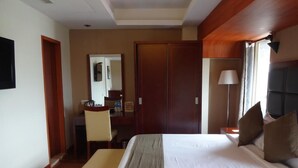 Une chambre d’hôtel avec un grand lit, un bureau avec une chaise, un miroir, une armoire et une fenêtre avec une lampe.