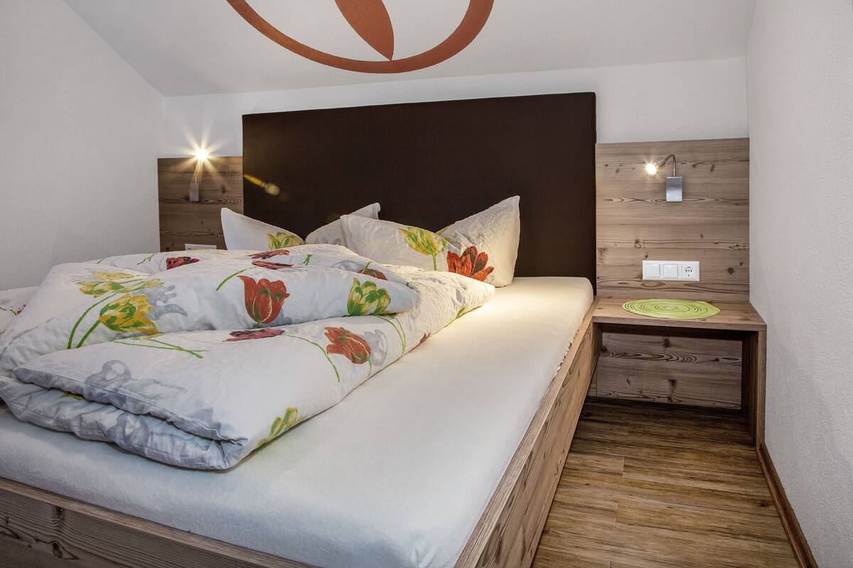 A neatly made bed with floral bedding, a wooden headboard, bedside lamps, and a nightstand with a power outlet.