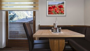 A dining area with a wooden table, black cushioned booth seating, a framed picture of red flowers, and a window with blinds. - Apart Bergland (Kaunertal)