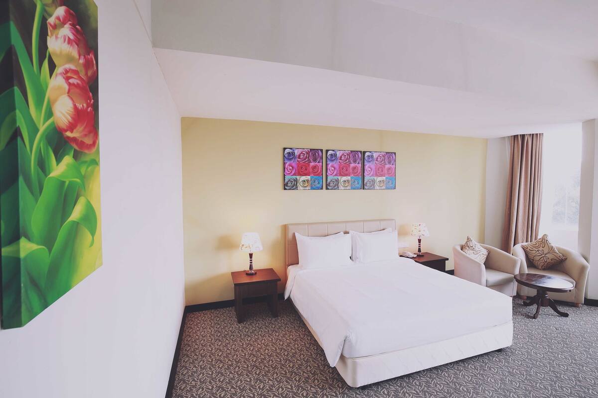A hotel room with a large bed, a nightstand with a lamp, a chair, a small table, and a wall with three framed artworks.