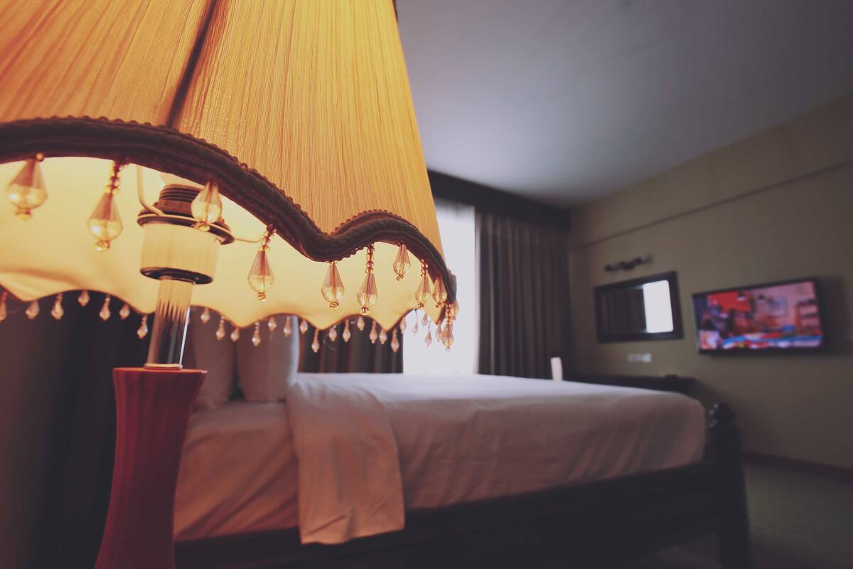 A hotel room with a bed, a large hanging lamp, and a television on the wall.