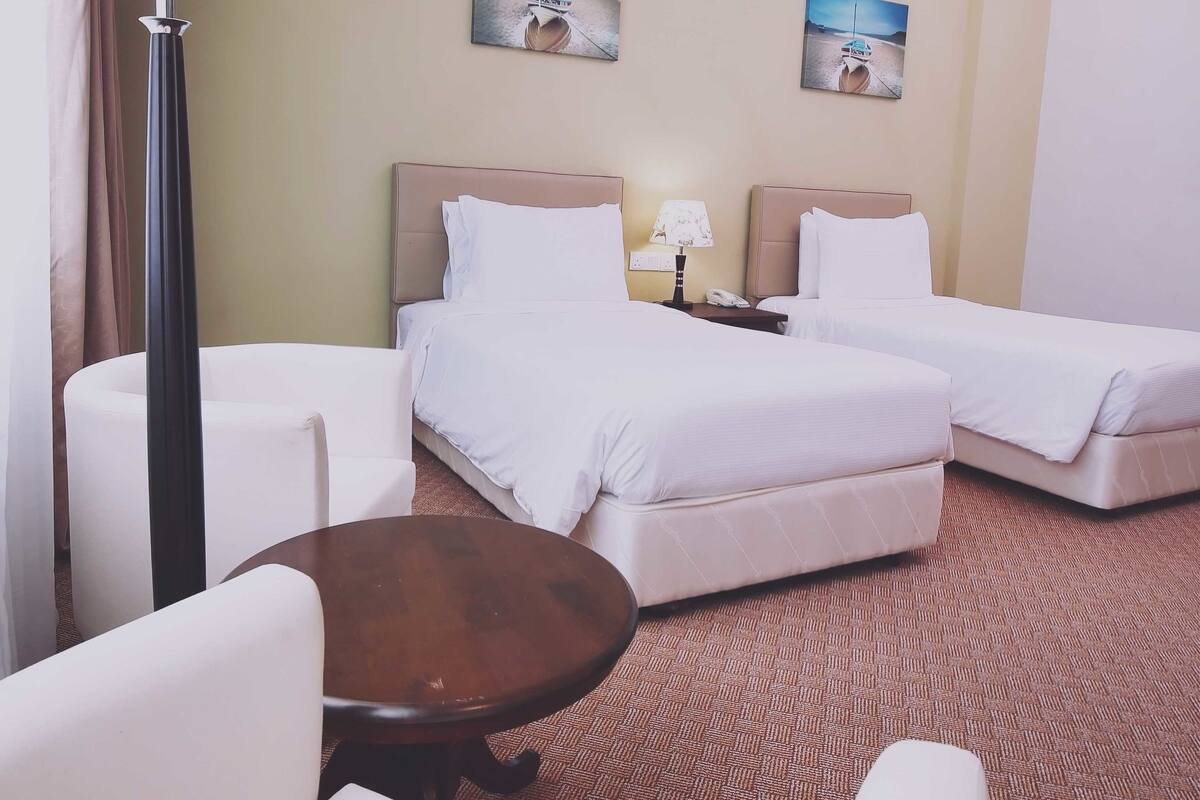 A hotel room with two beds, a desk, a chair, a lamp, and two wall-mounted paintings.