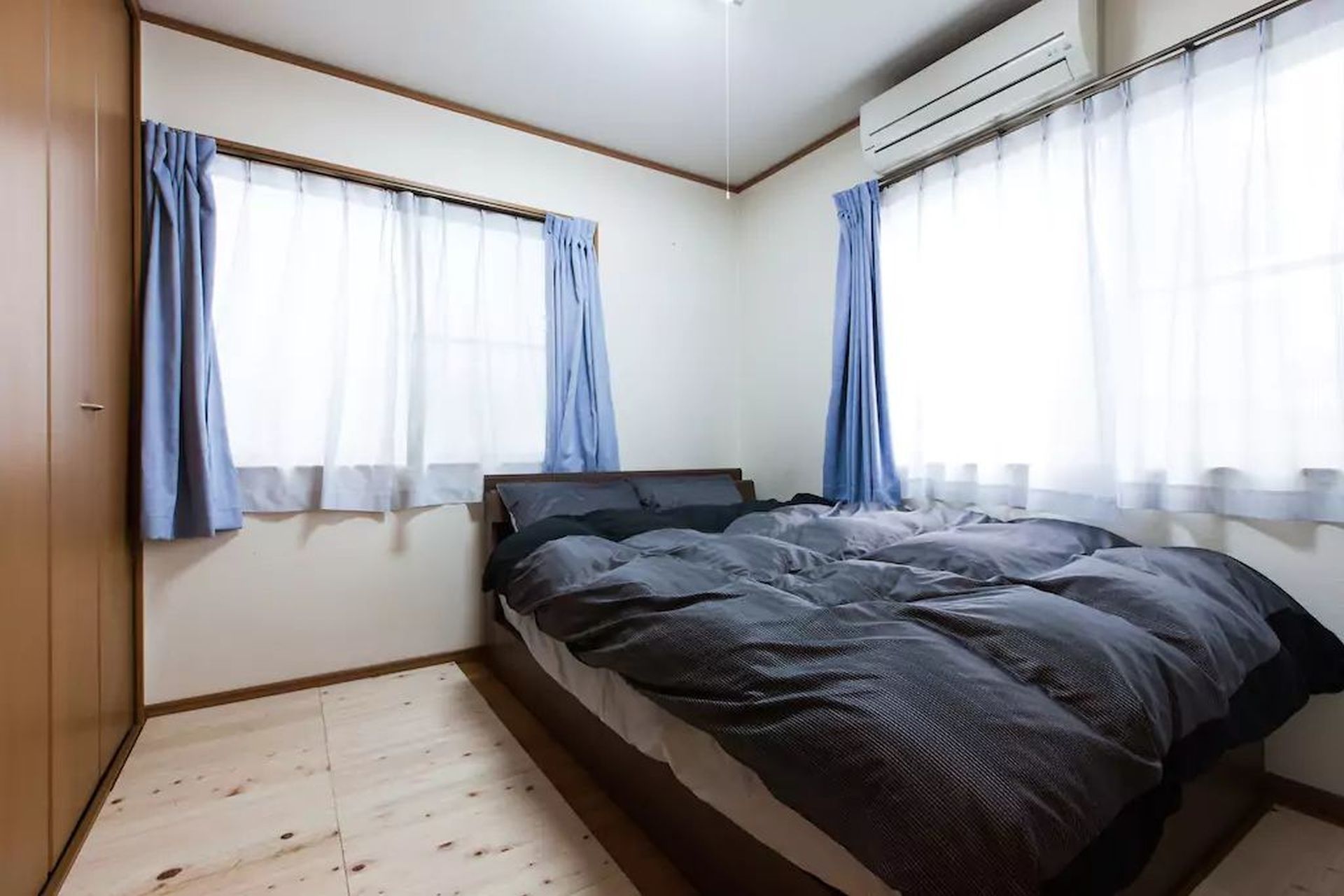 A bedroom with a bed, two windows with curtains, an air conditioning unit, and a wooden floor.