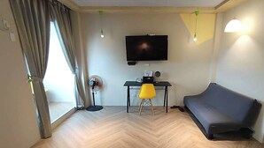 A living room with a flat-screen TV mounted on the wall, a desk with a chair, a sofa, and a standing fan. - Light Shadow (Budai)