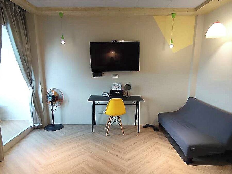 A living room with a flat-screen TV mounted on the wall, a desk with a chair, a sofa, and a standing fan.