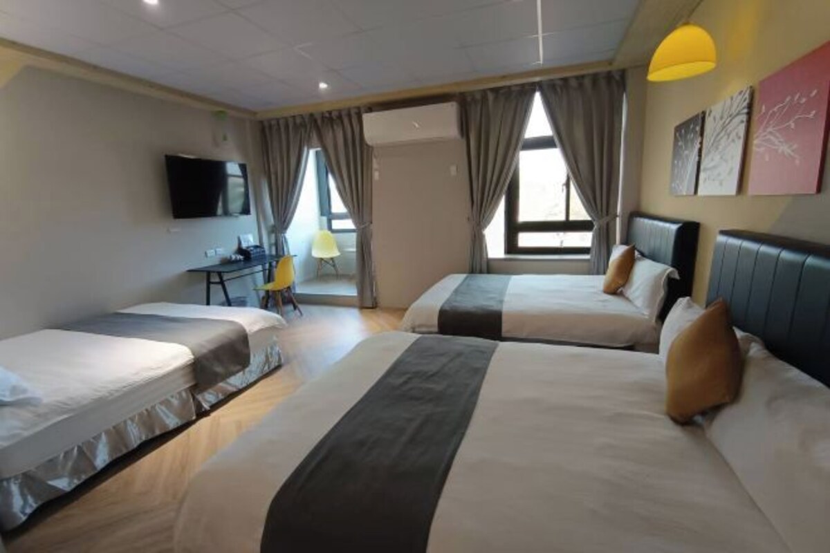 A hotel room with two beds, a desk, a chair, a TV, and large windows with curtains.
