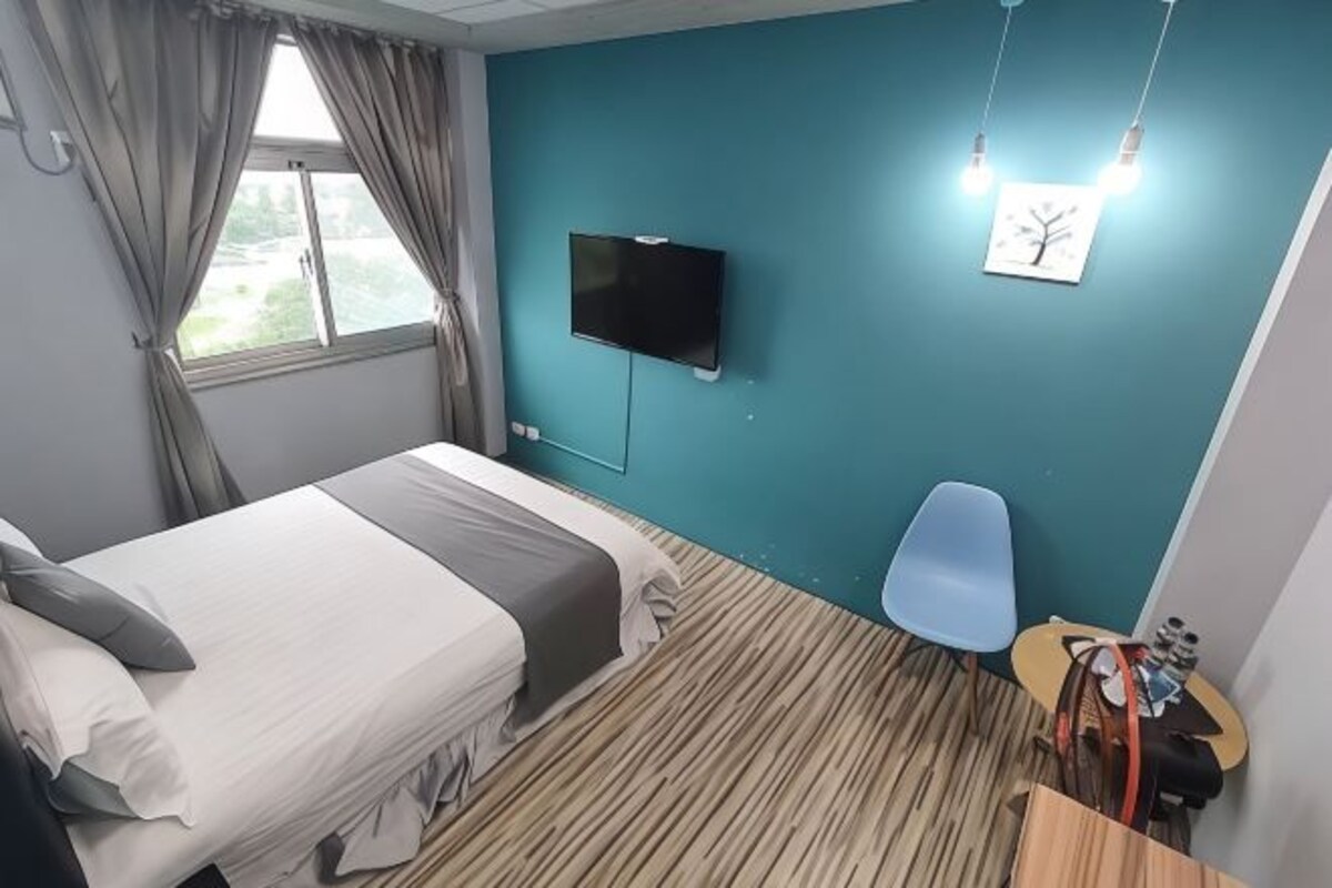 A hotel room with a bed, a TV mounted on the wall, a chair, and a small table with items on it.