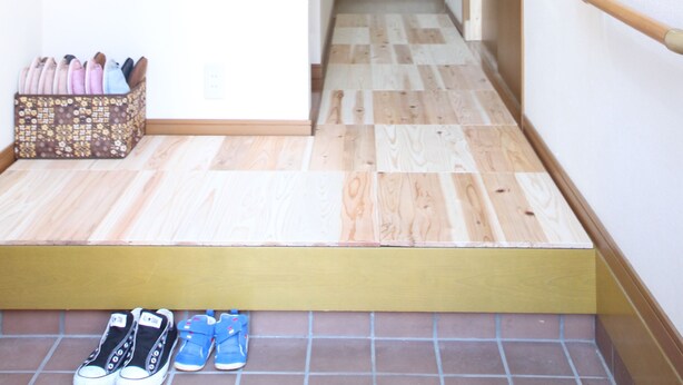 A narrow hallway with wooden flooring and a small step leading to a tiled area. A shoe rack with various shoes is visible on the left.