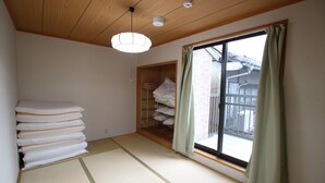 A room with tatami flooring, a stack of white pillows, a wooden shelf, and a sliding glass door with a view of the outside.