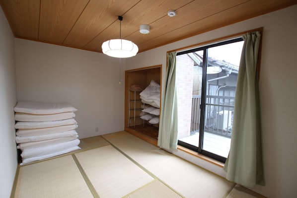 A room with tatami flooring, a stack of white pillows, a wooden shelf, and a sliding glass door with a view of the outside.