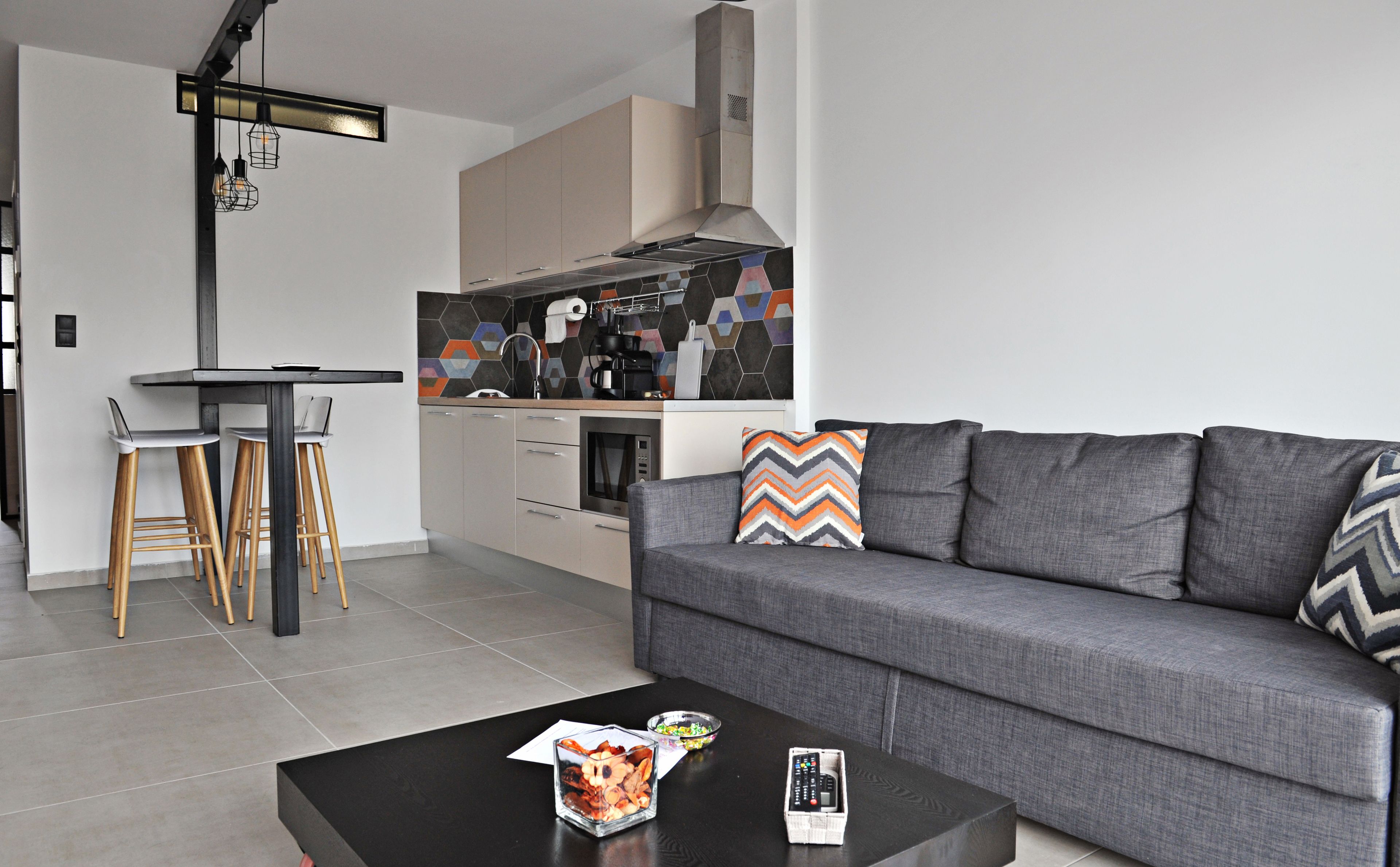 A modern living room with a grey sofa, wooden bar stools, a coffee table, and a kitchen in the background.
