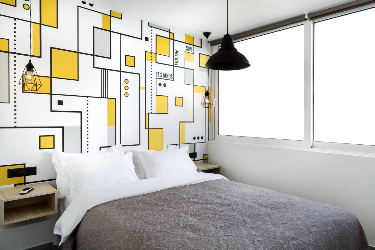 A modern bedroom with a geometric-patterned wall, a bed with white and gray bedding, a bedside table with a lamp, and a window with a view.