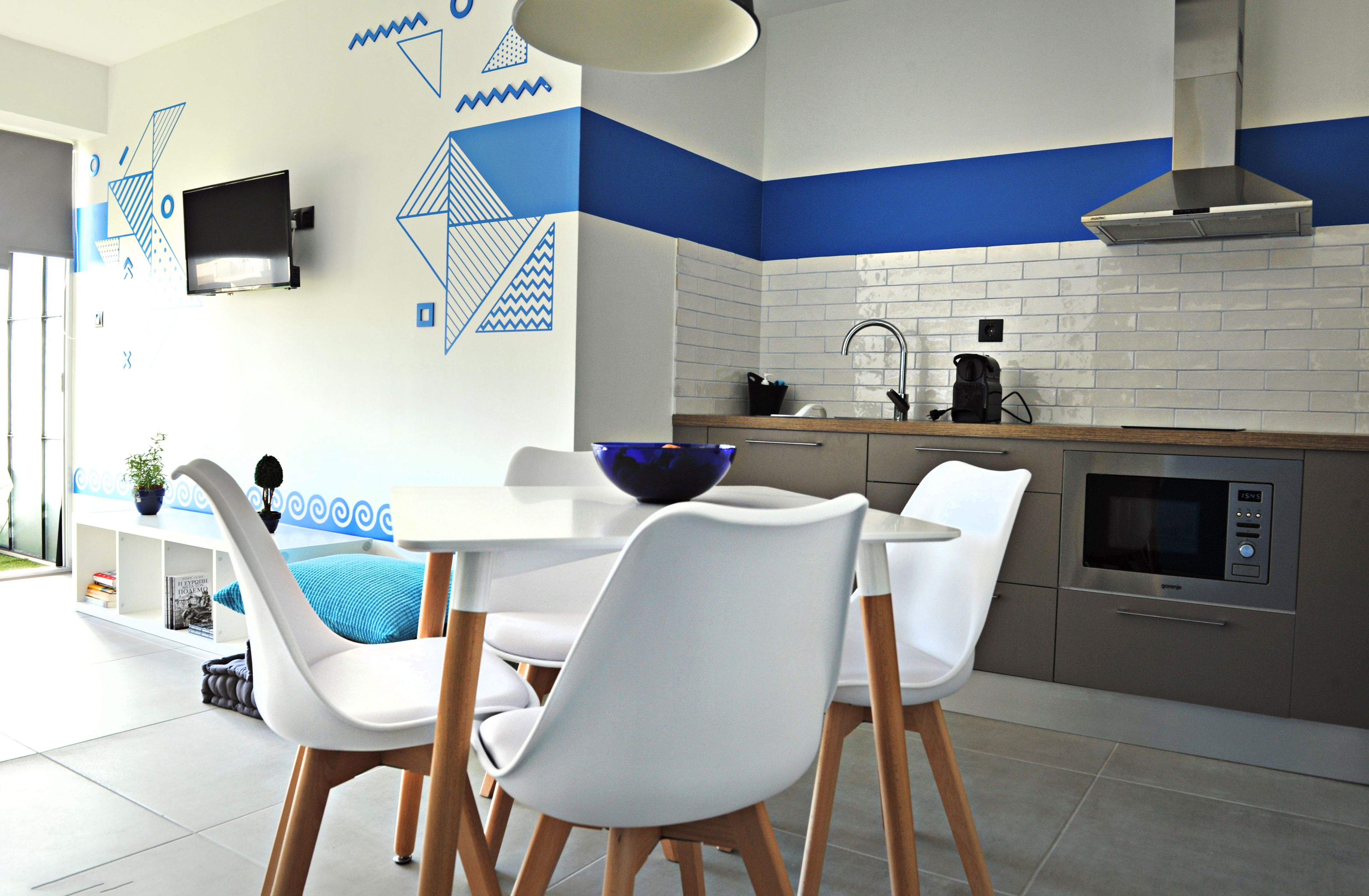 Una cocina moderna con diseños geométricos blancos y azules en las paredes, una mesa de comedor blanca con sillas y electrodomésticos de acero inoxidable.