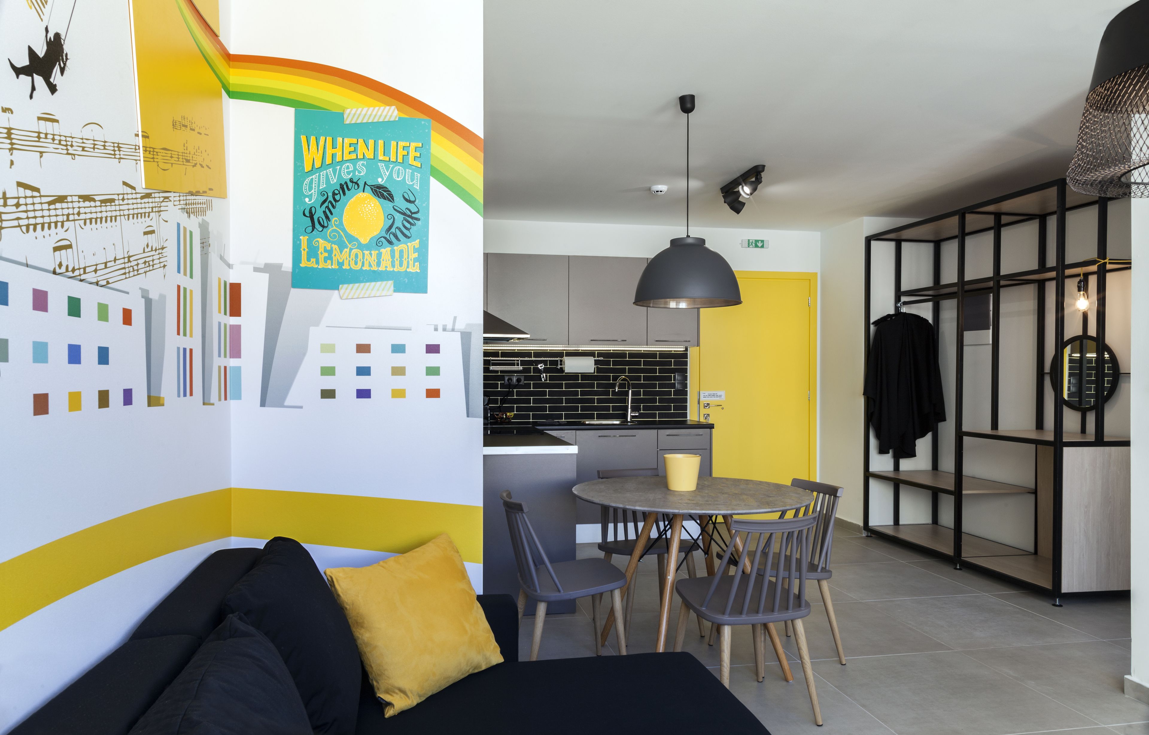 Eine moderne Küche mit gelber Tür, ein Essbereich mit rundem Tisch und Stühlen sowie ein schwarzes Sofa mit gelbem Kissen.