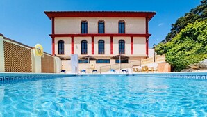 Un hotel con piscina, fachada roja y blanca, y vistas al entorno verde.