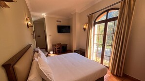 Habitación de hotel con una cama grande, un escritorio, una silla, un televisor y un balcón con cortinas.