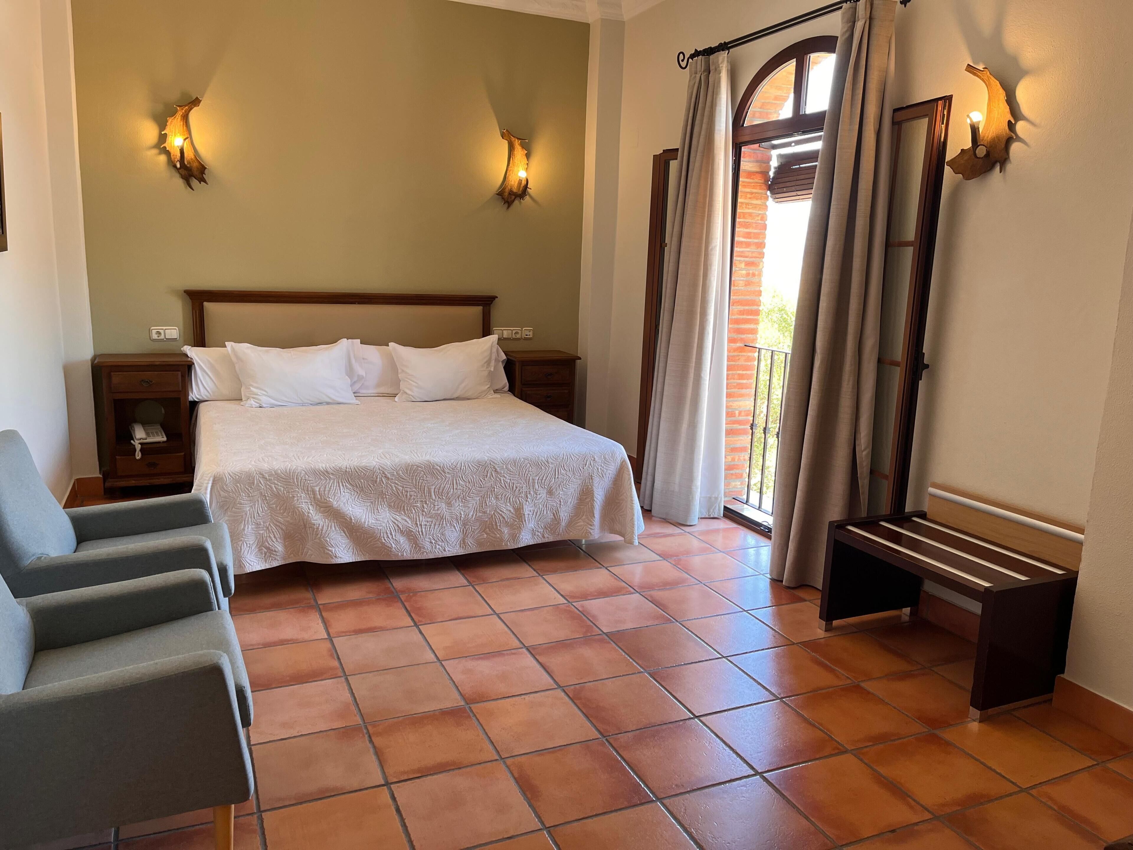Habitación de hotel con cama, dos lámparas de noche, mesita de noche, una silla y un balcón con cortinas.