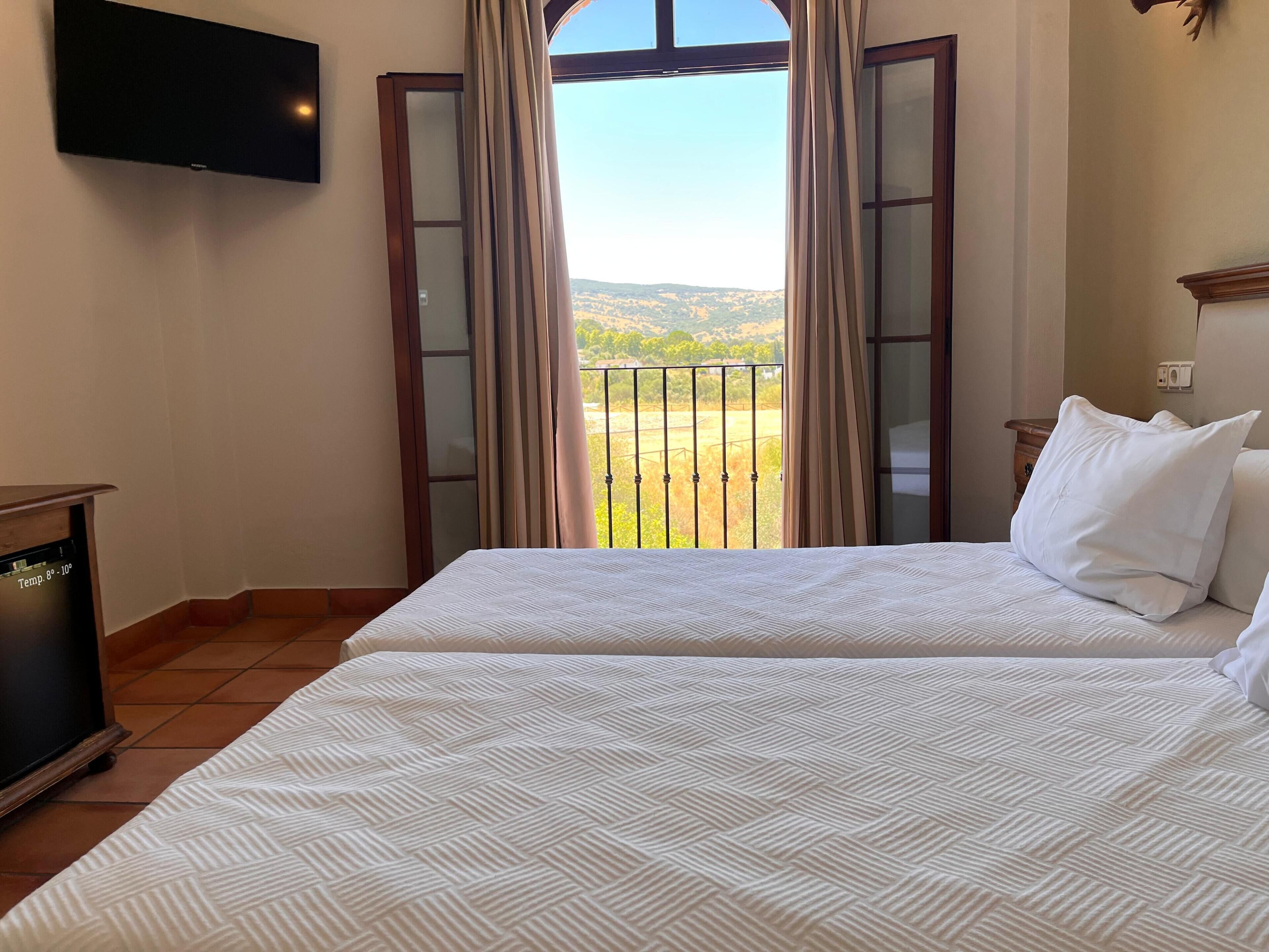 Una habitación de hotel con cama, televisión montada en la pared y balcón con cortinas.